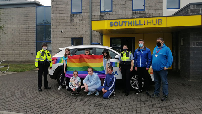 Group of adults and children outside building holding rainbow flag. Building sign, grey on yellow, reading, Southill Hub